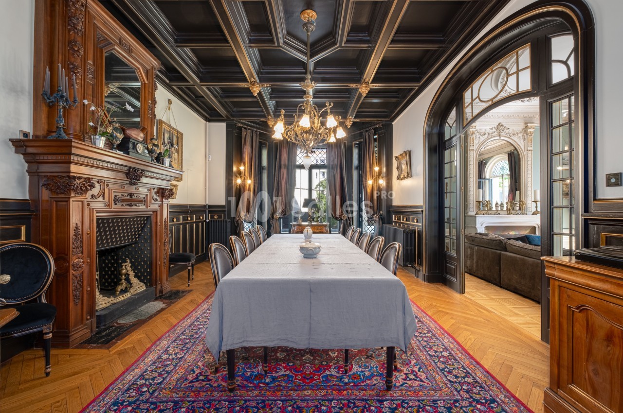 Salle à manger élégante avec table dressée, cheminée en bois sculpté, lustre et parquet en chevrons.