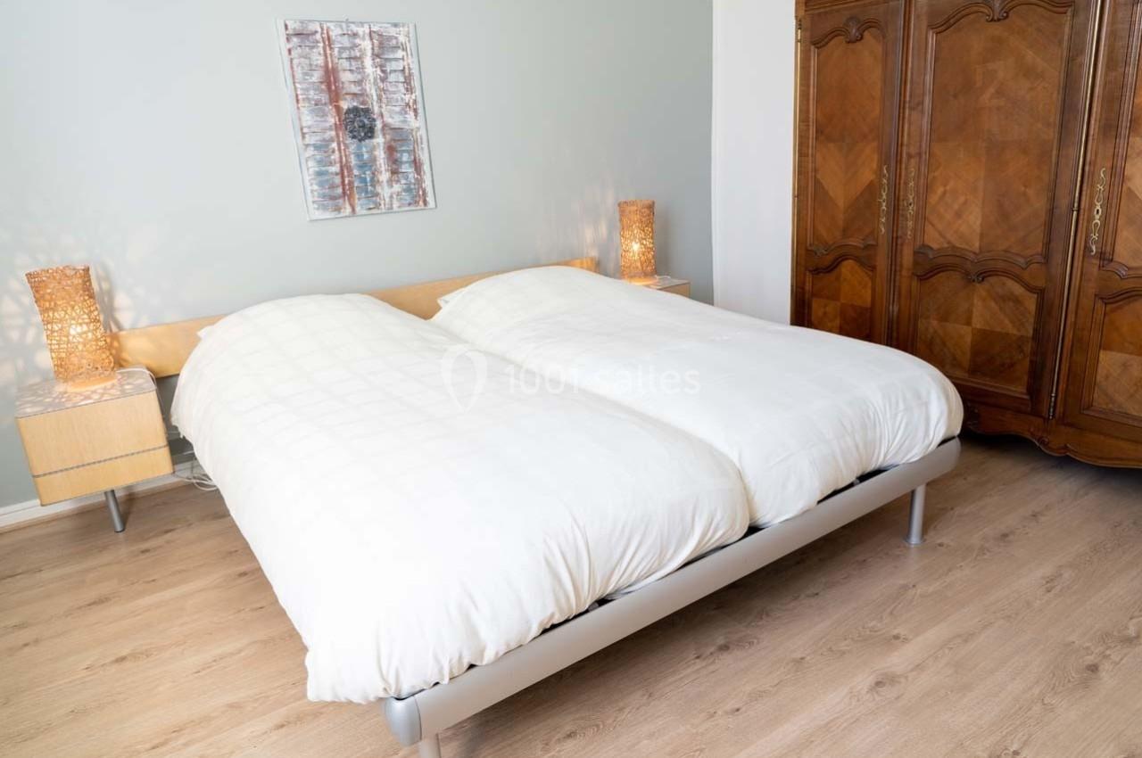 Lit double avec couette blanche dans une chambre lumineuse, encadré par deux tables de chevet et une armoire en bois.