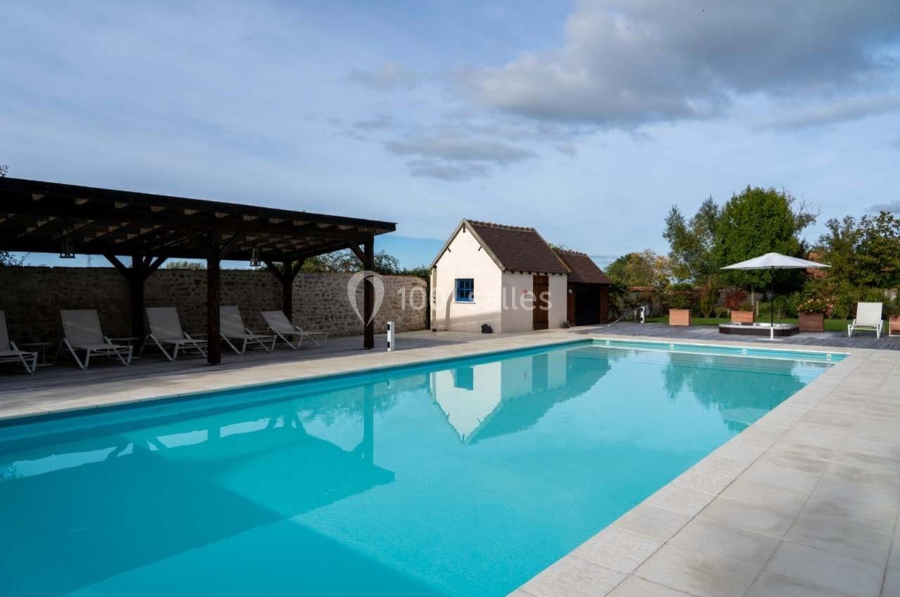 Piscine extérieure avec terrasse en pierre, chaises longues sous un abri et petite maison en arrière-plan.