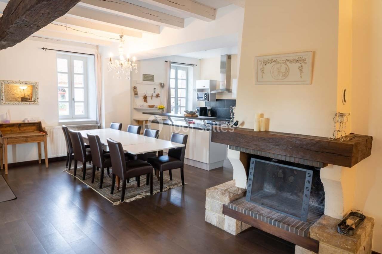 Salle à manger lumineuse avec table et chaises en bois sombre, cheminée en pierre et cuisine ouverte en arrière-plan.