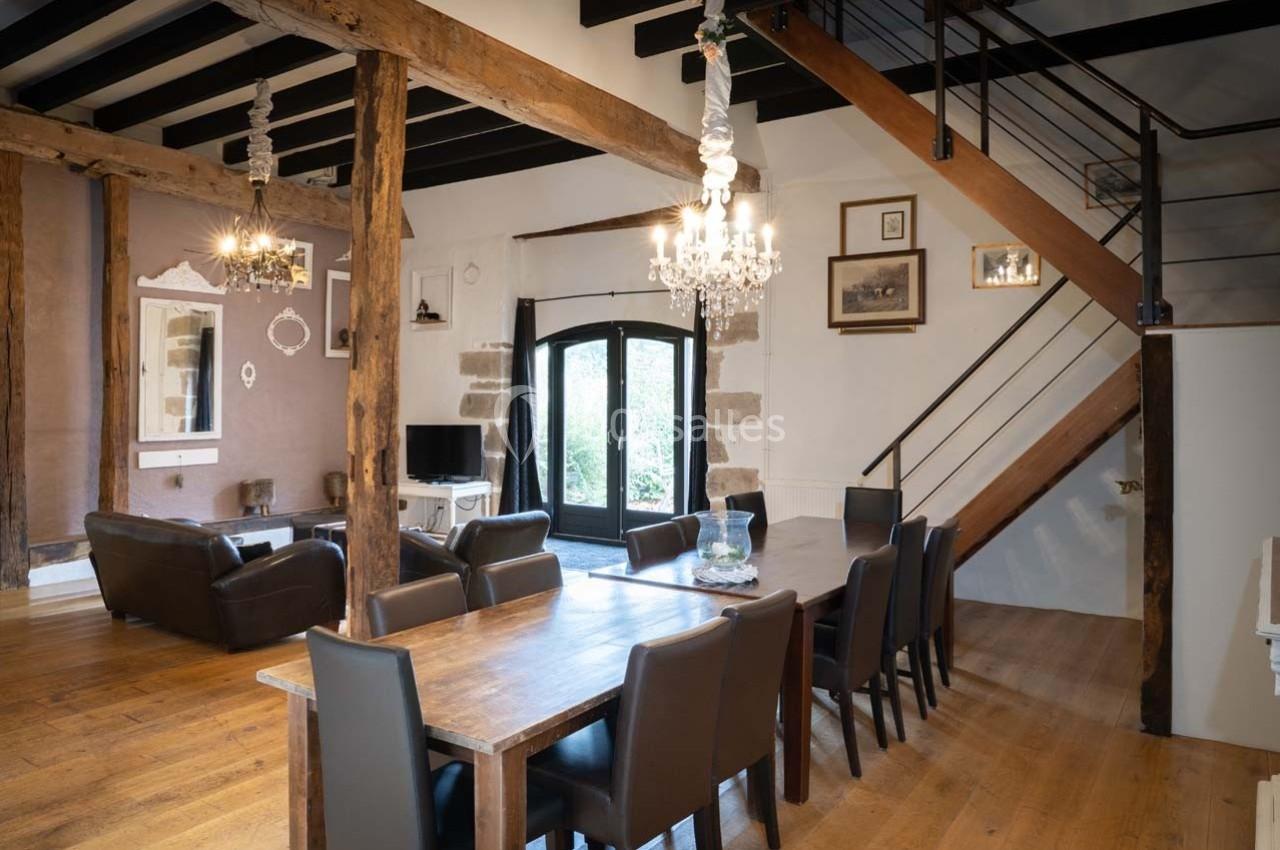 Salle à manger spacieuse avec table en bois, chaises noires, poutres apparentes et escalier en métal.