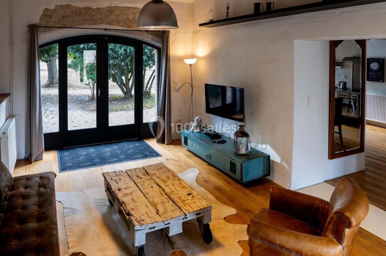 Salon lumineux avec canapé en cuir, table basse en bois, télévision, et grande baie vitrée donnant sur un jardin.