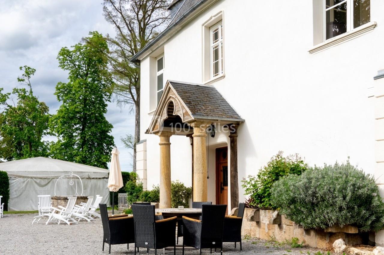 Façade d'une maison blanche avec une entrée voûtée, entourée de mobilier de jardin et de végétation.