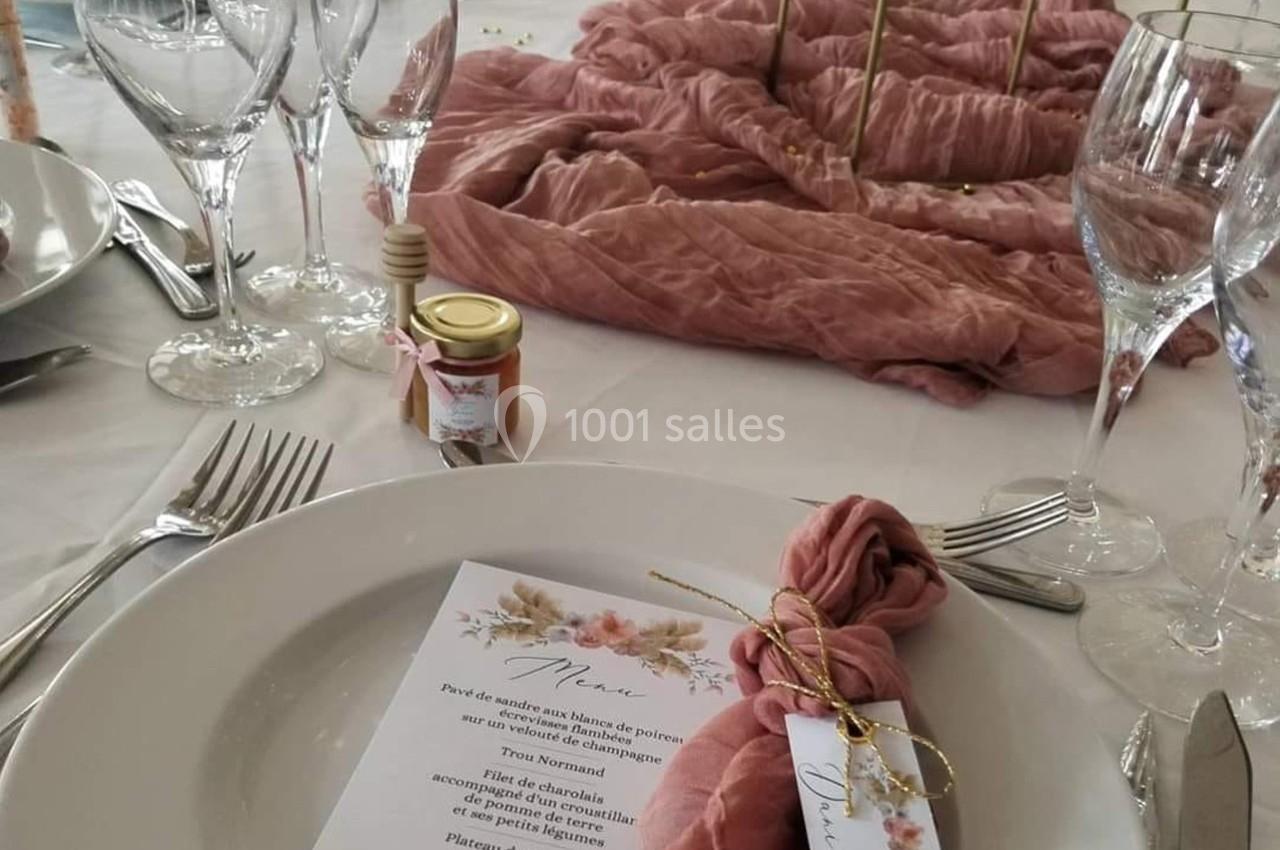 Table dressée avec une assiette contenant un menu floral, une serviette rose nouée et un pot de miel en décoration.