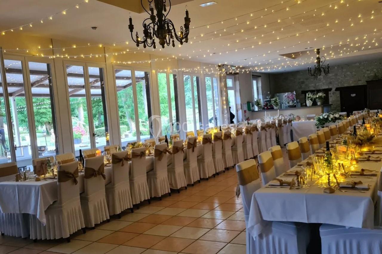 Salle de réception décorée pour un événement, avec longues tables dressées, chaises ornées et guirlandes lumineuses…