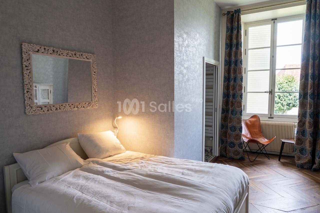 Chambre lumineuse avec lit double, miroir mural, lampe de chevet allumée, chaise en cuir et fenêtre donnant sur l'extérieur.