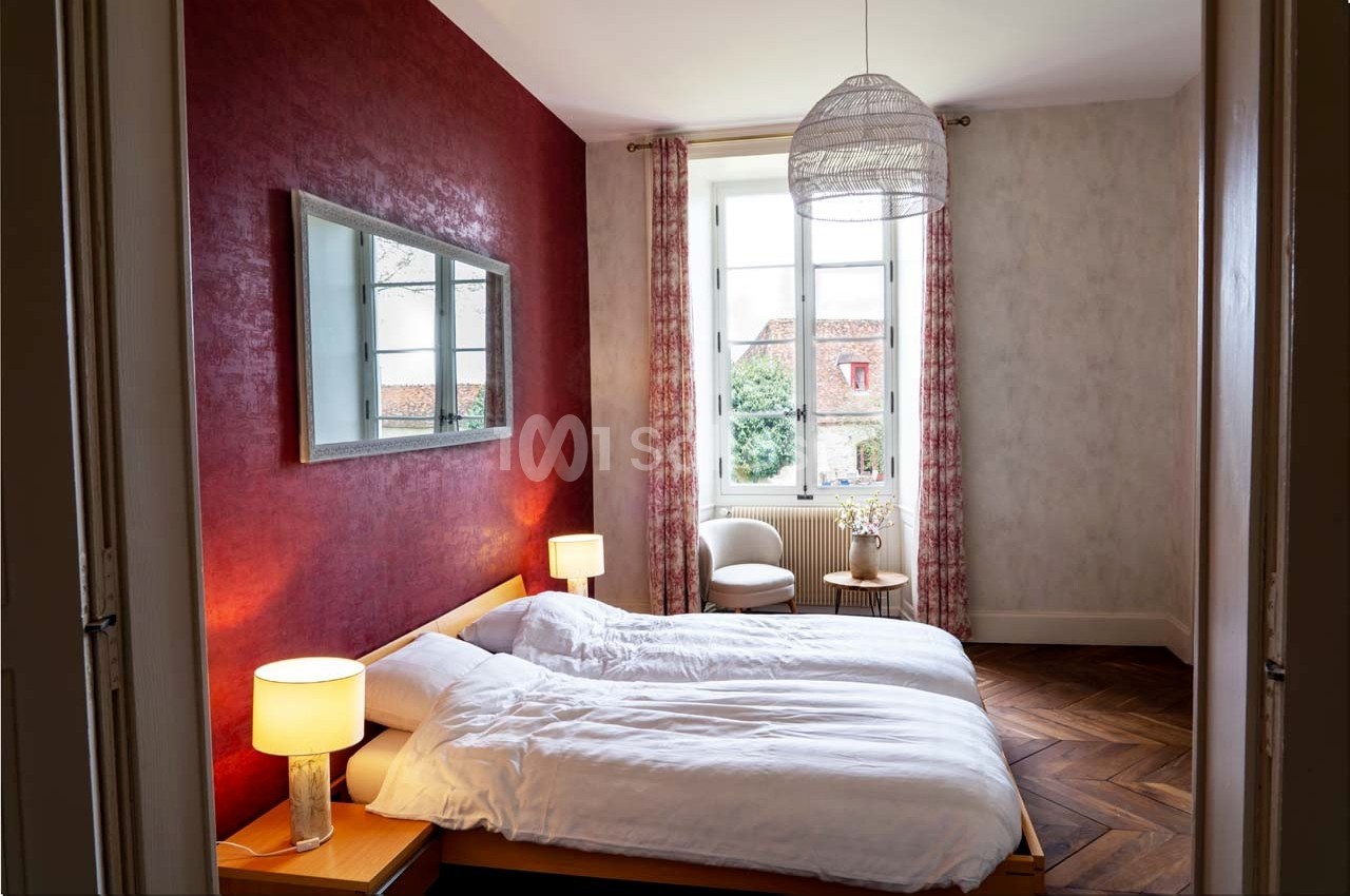 Chambre lumineuse avec deux lits, murs rouges et gris, parquet en bois, grande fenêtre et lampe suspendue.