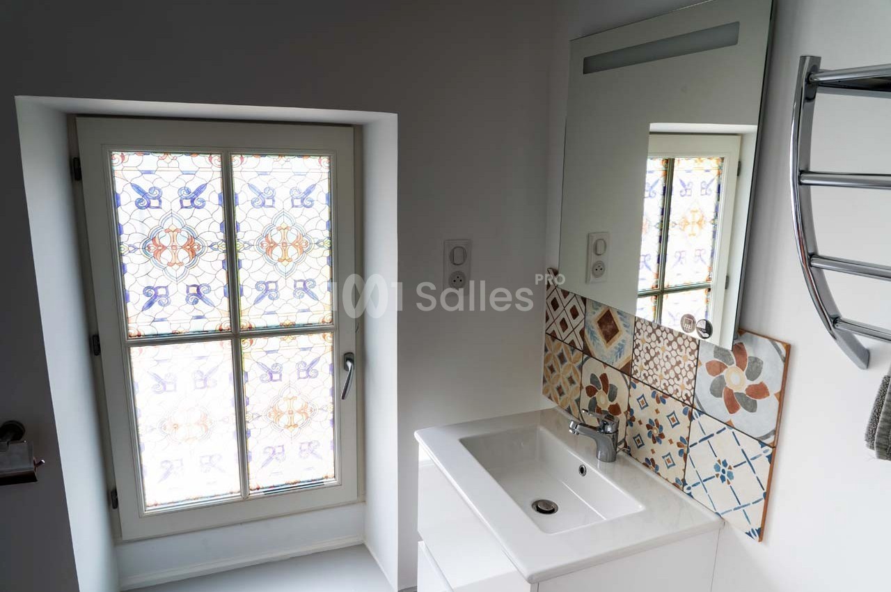 Lavabo blanc avec miroir, carrelage décoratif en mosaïque et fenêtre à motifs colorés dans une salle de bain lumineuse.