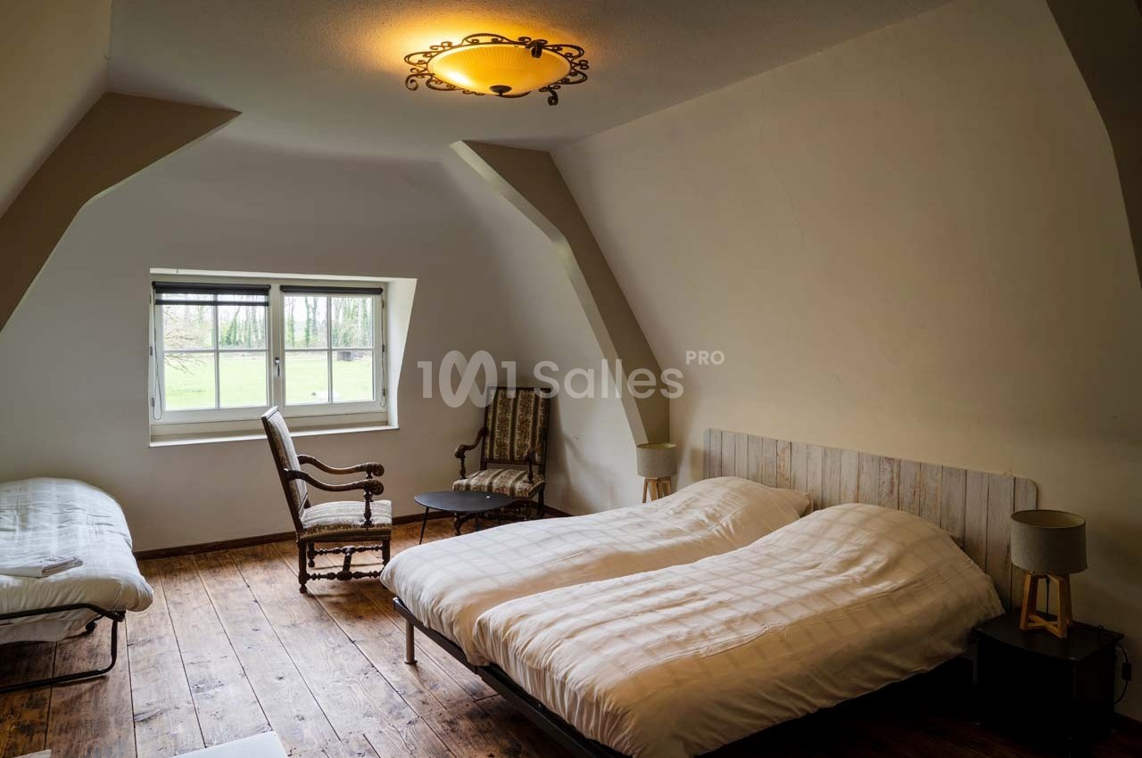 Chambre mansardée avec deux lits, un fauteuil, une chaise, un lit d'appoint et une fenêtre donnant sur un paysage verdoyant.