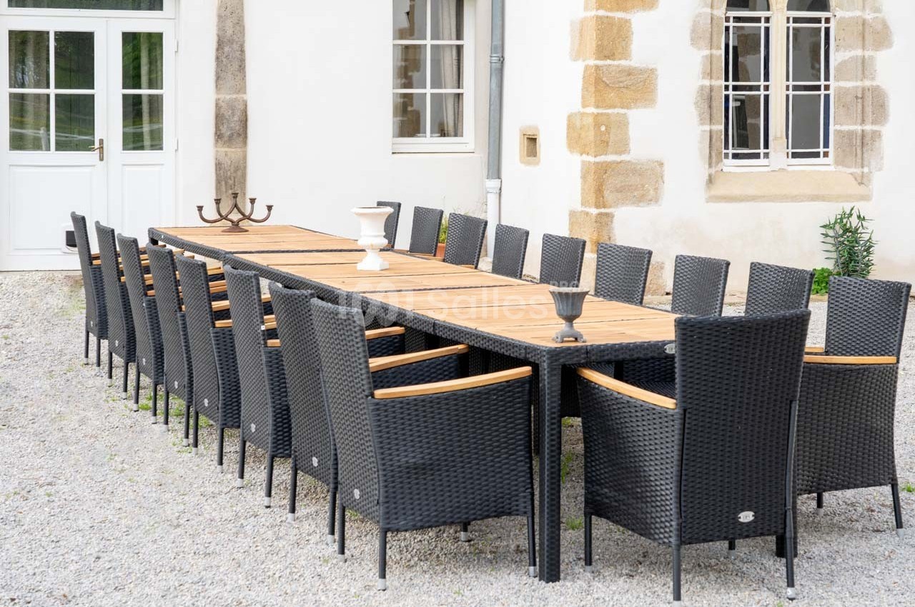 Grande table en bois entourée de chaises en rotin noir, installée sur un sol en gravier devant un bâtiment clair.
