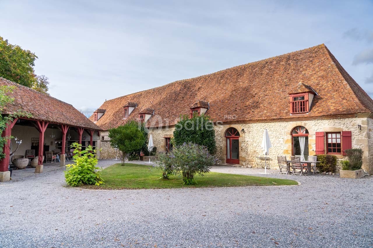 Bâtisse en pierre avec toit en tuiles, entourée de végétation et d'une cour gravillonnée, sous un ciel légèrement nuageux.