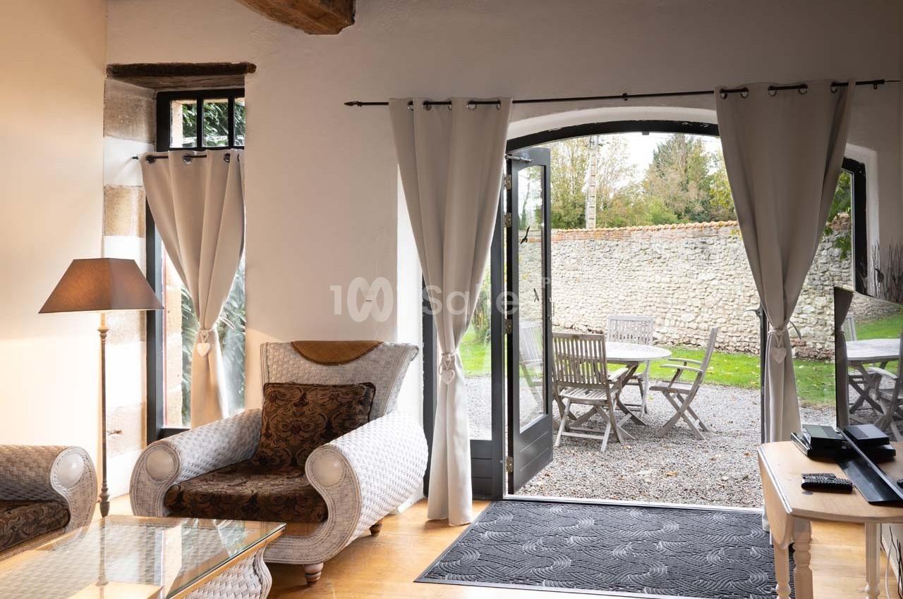 Salon lumineux avec fauteuils, table basse et porte-fenêtre ouverte sur une terrasse avec table et chaises en bois.