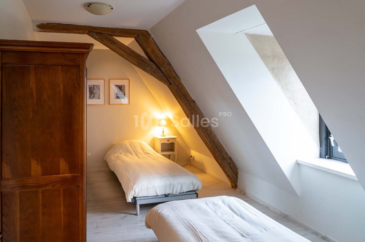 Chambre mansardée avec deux lits simples, une poutre apparente, une lampe de chevet et une fenêtre inclinée.