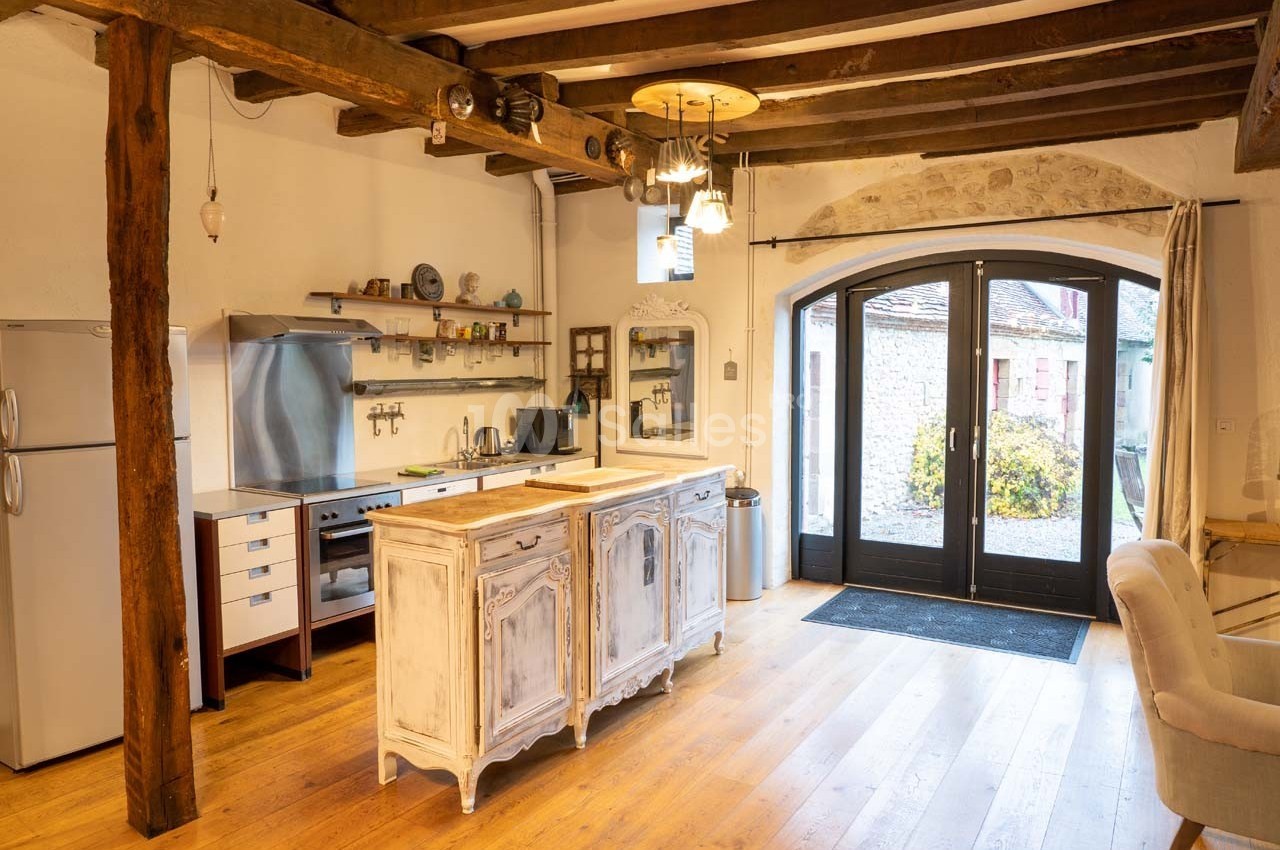 Cuisine rustique avec poutres apparentes, meubles en bois patiné, sol en parquet et porte-fenêtre donnant sur une cour.