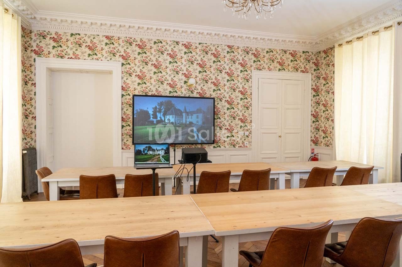 Salle de réunion lumineuse avec des tables en bois, des chaises en cuir, un écran et un décor floral sur les murs.