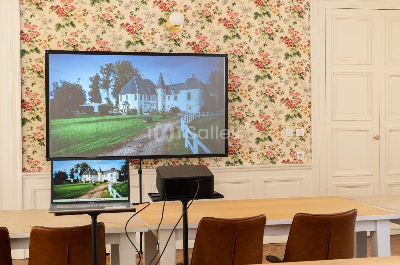 Écran projetant l'image d'un château, installé dans une salle avec des tables, des chaises et un mur à motif floral.