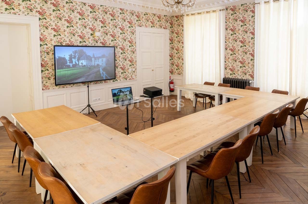 Salle de réunion lumineuse avec tables en U, chaises en cuir, écran de présentation et décoration murale florale.