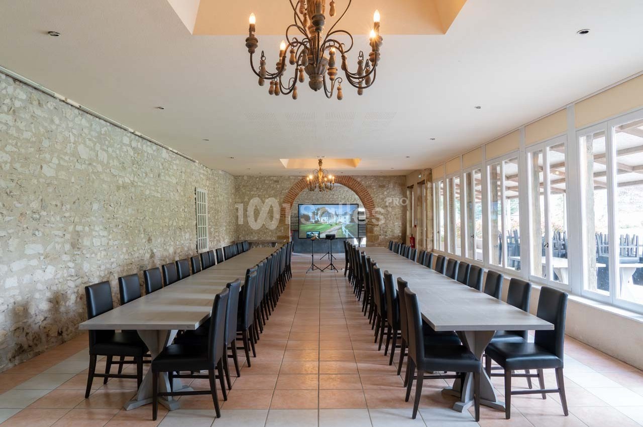 Salle de réception lumineuse avec murs en pierre, grandes tables alignées, chaises noires et lustres suspendus.