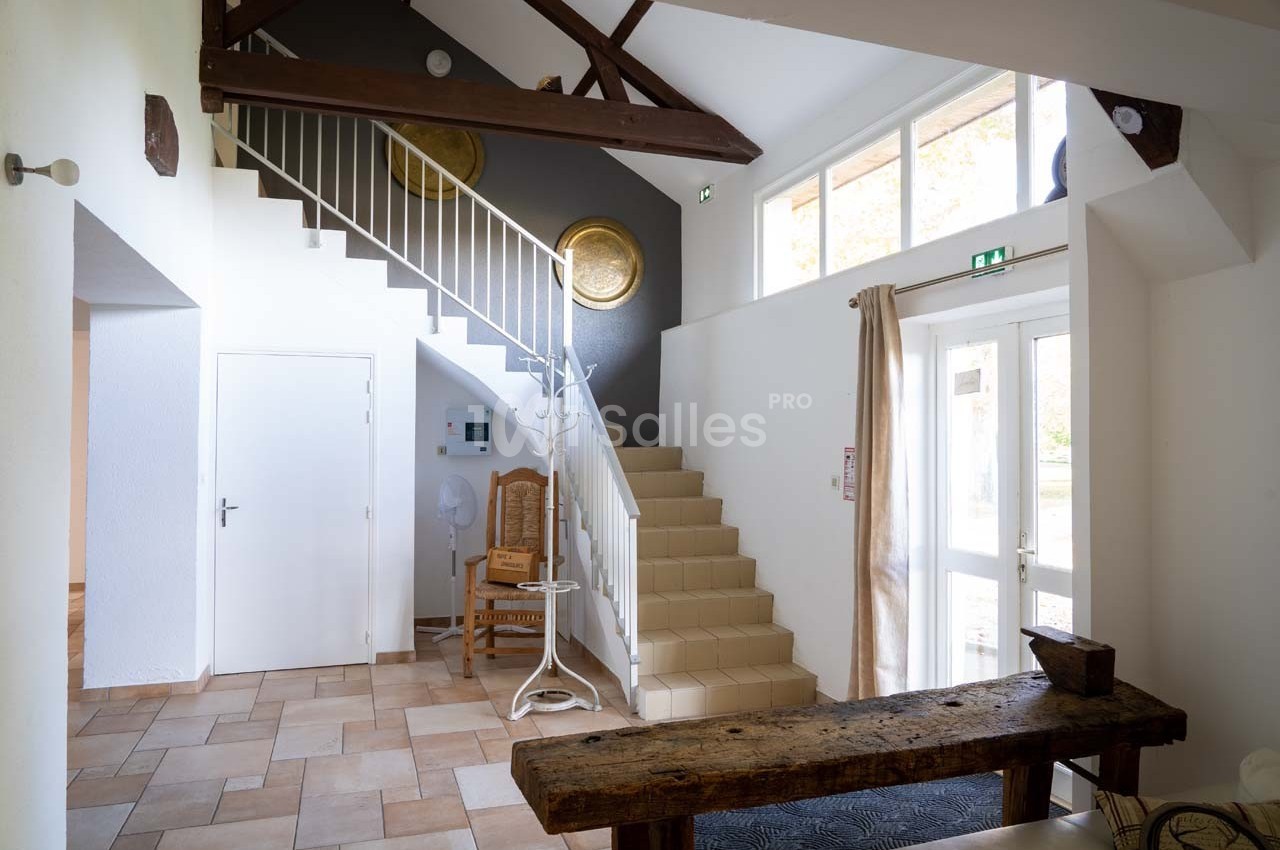 Entrée lumineuse avec escalier en carrelage beige, poutres apparentes et mobilier en bois rustique.
