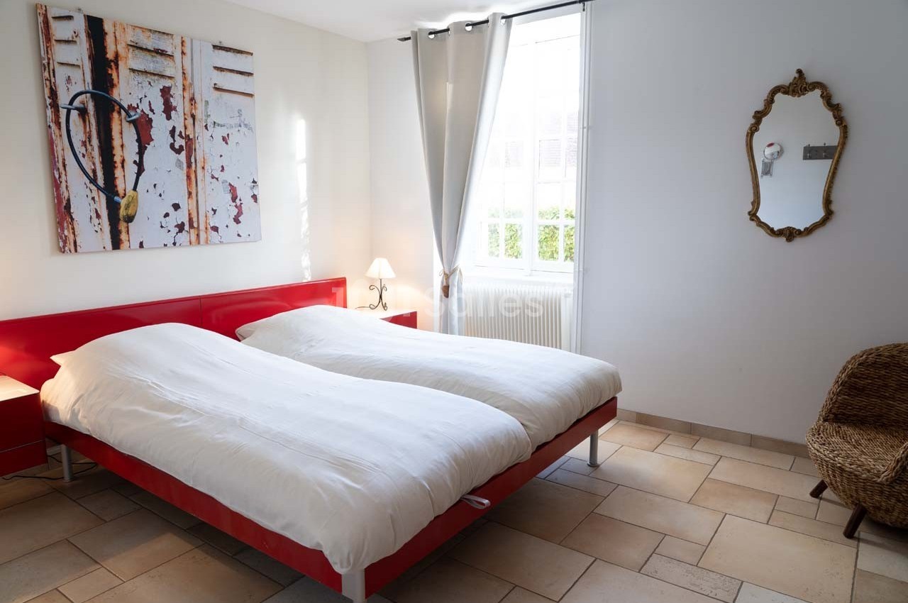 Chambre lumineuse avec deux lits simples, un miroir mural, un fauteuil en osier et un tableau décoratif.