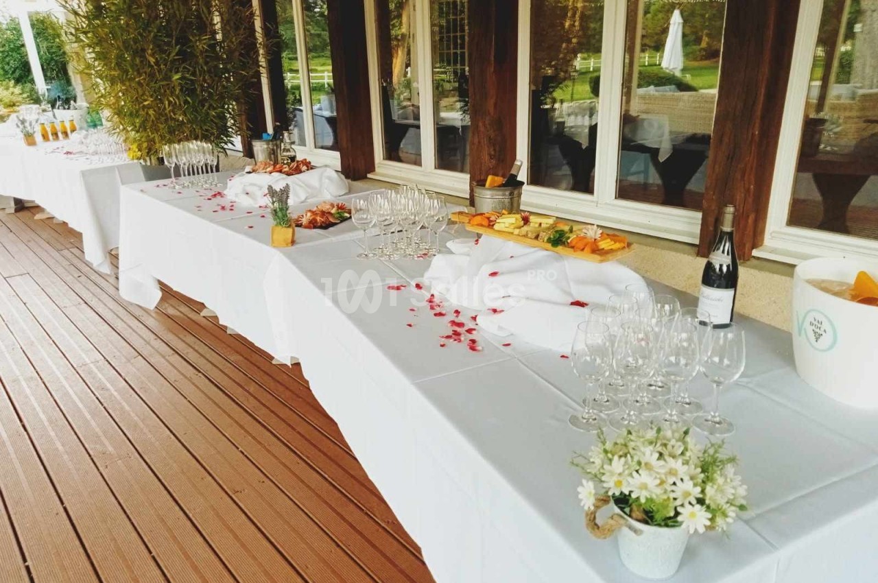 Tables dressées avec nappes blanches, verres, bouteilles et plats variés sur une terrasse en bois près de grandes fenêtres.