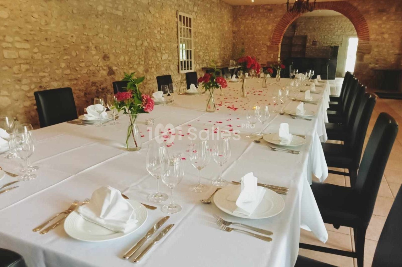Grande table rectangulaire dressée avec nappes blanches, vaisselle élégante et décorations florales dans une salle en pierre.