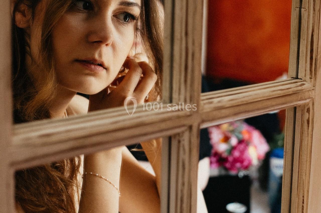 Femme ajustant une boucle d'oreille devant une fenêtre, avec un bouquet de fleurs en arrière-plan.