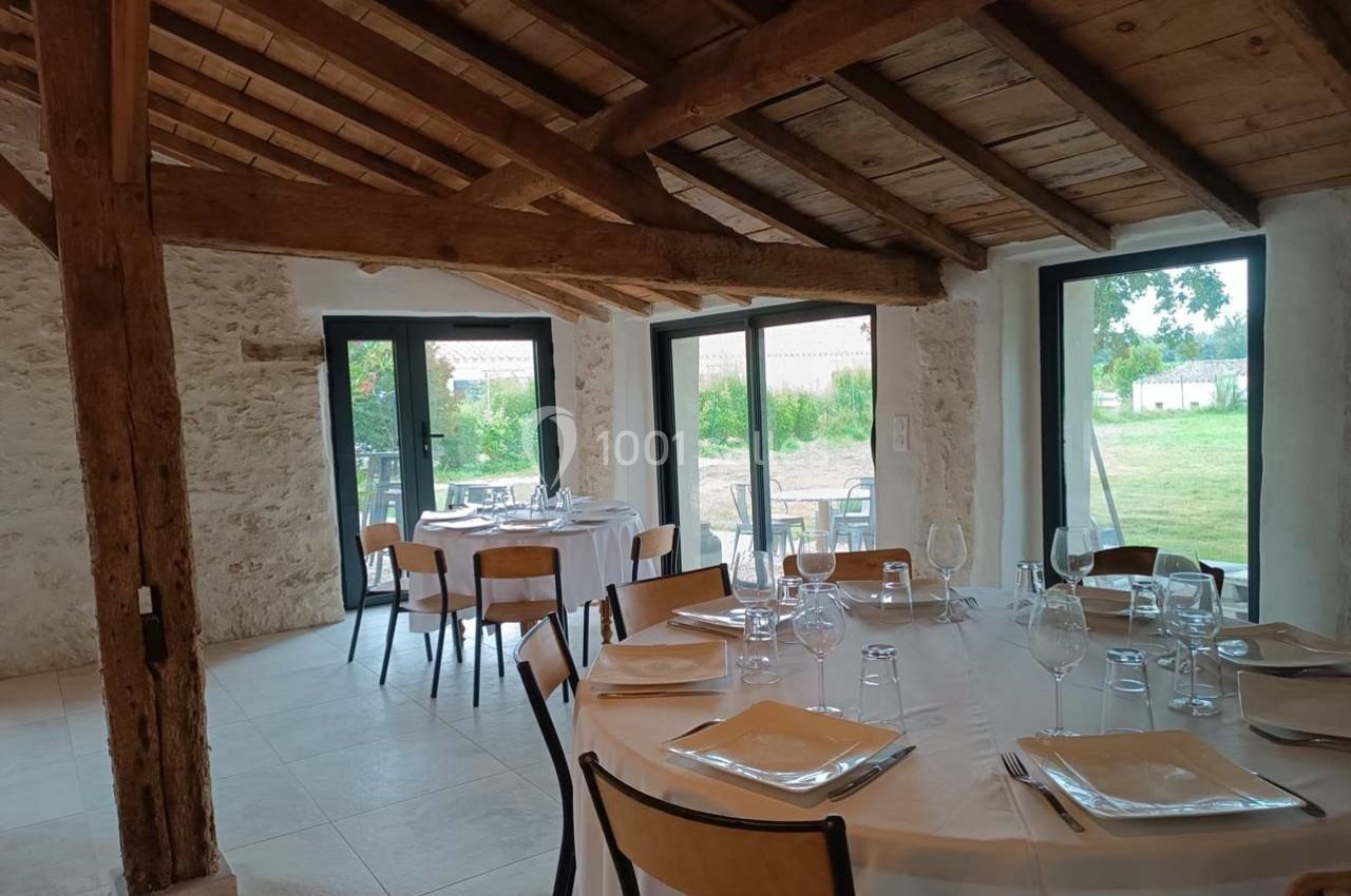 Salle de réception lumineuse avec tables dressées, grandes baies vitrées et poutres apparentes en bois.