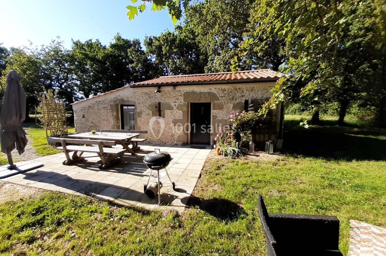 Maison en pierre avec terrasse aménagée, table de pique-nique, barbecue et jardin verdoyant sous un ciel ensoleillé.