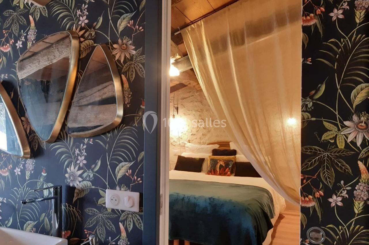 Chambre cosy avec lit double, rideaux légers, murs en pierre et papier peint floral, vue partielle sur une salle de bain.