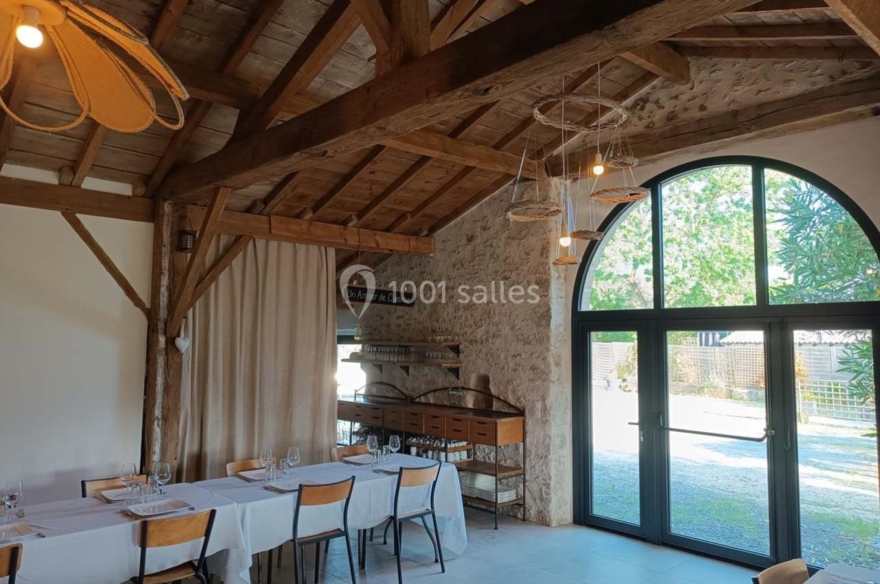 Salle lumineuse avec poutres apparentes, tables dressées et grande baie vitrée donnant sur un extérieur verdoyant.
