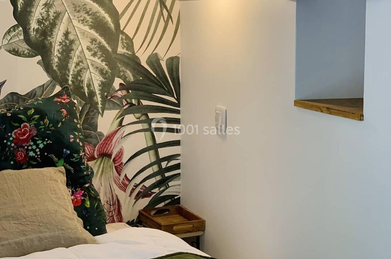 Mur décoré de papier peint tropical, lit avec coussins colorés, étagère en bois et lampe murale.