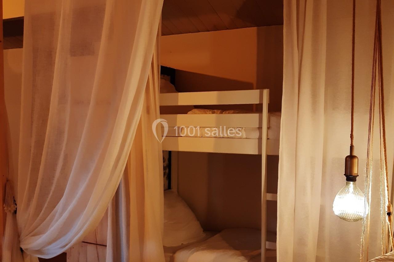 Lit superposé blanc derrière des rideaux légers dans une chambre chaleureuse éclairée par une lampe suspendue.
