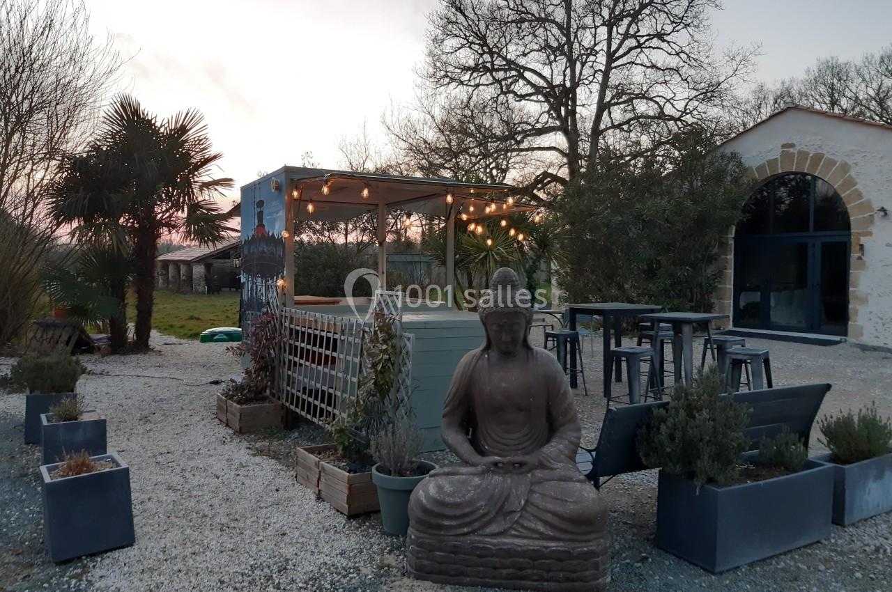 Statue de Bouddha devant une terrasse éclairée avec des guirlandes, entourée de plantes et de mobilier extérieur.