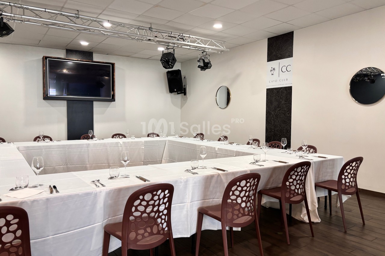 Salle de réunion avec tables disposées en U, nappes blanches, chaises marron et écran mural.
