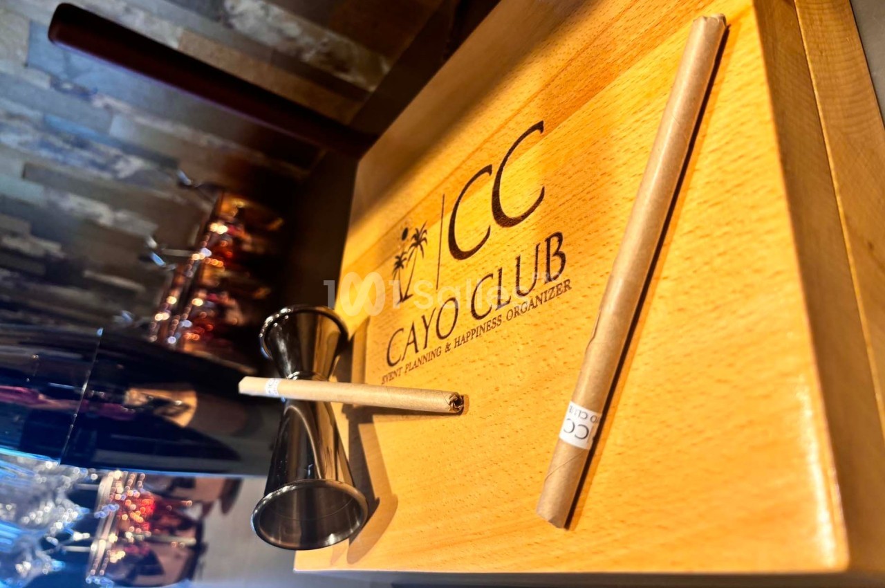 Planche en bois gravée ’Cayo Club’ avec un doseur en métal et deux crayons posés dessus, en arrière-plan des verres.