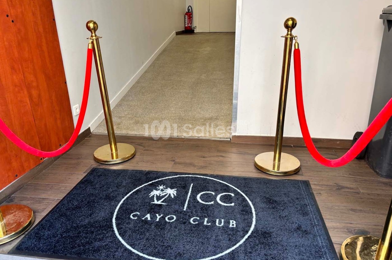 Tapis noir marqué ’Cayo Club’ devant une entrée délimitée par des poteaux dorés et des cordons rouges.