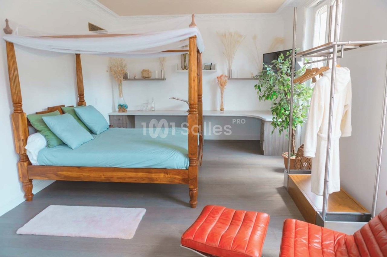 Chambre lumineuse avec lit à baldaquin en bois, bureau intégré, portant à vêtements et fauteuils en cuir rouge.