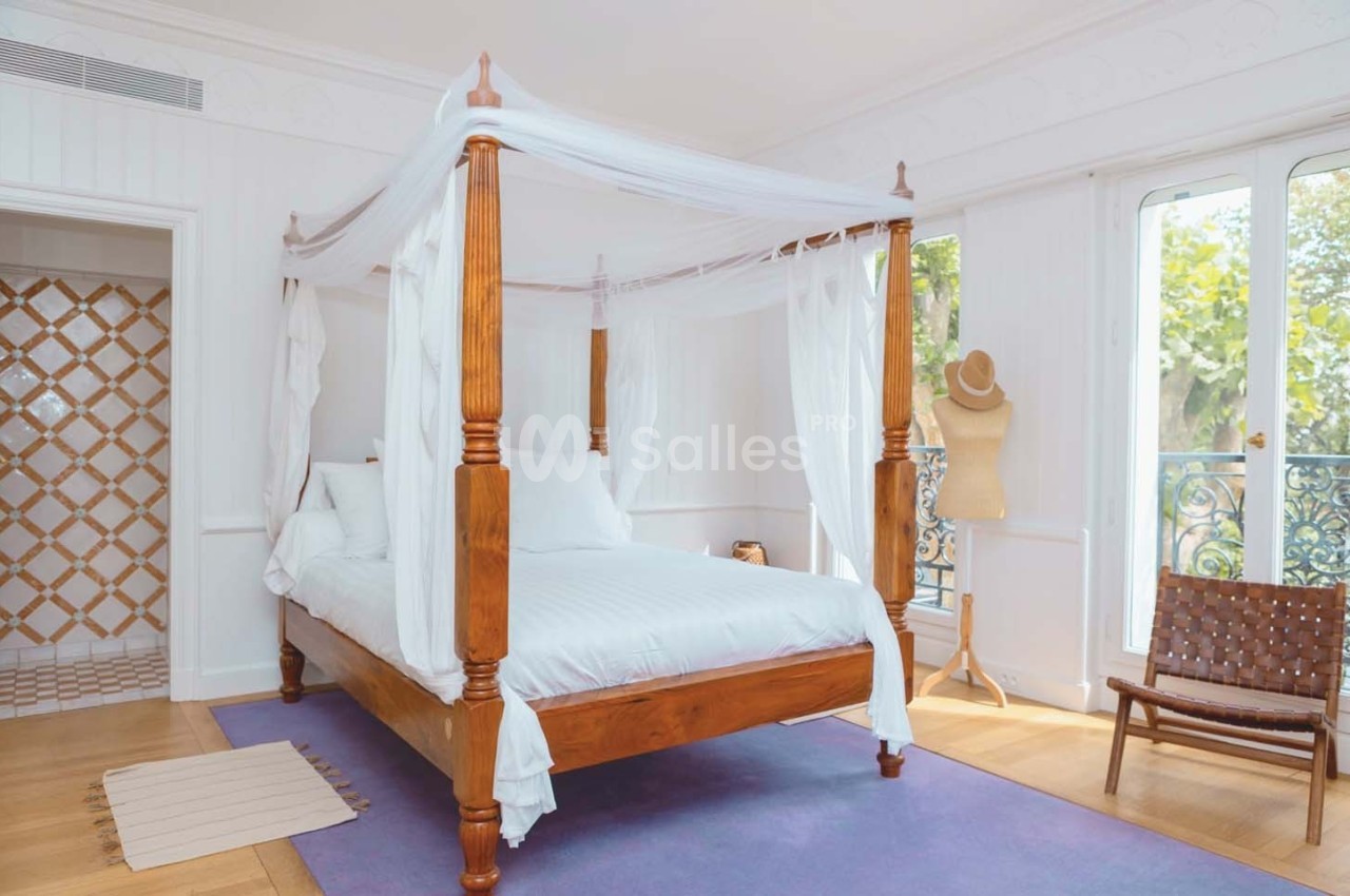 Lit à baldaquin en bois avec voilages blancs dans une chambre lumineuse, parquet clair et vue sur un balcon.