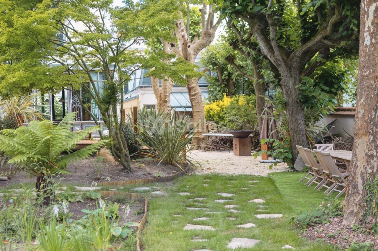 Jardin aménagé avec pelouse, allée en pierres, arbres, plantes variées, mobilier de jardin et barbecue en arrière-plan.