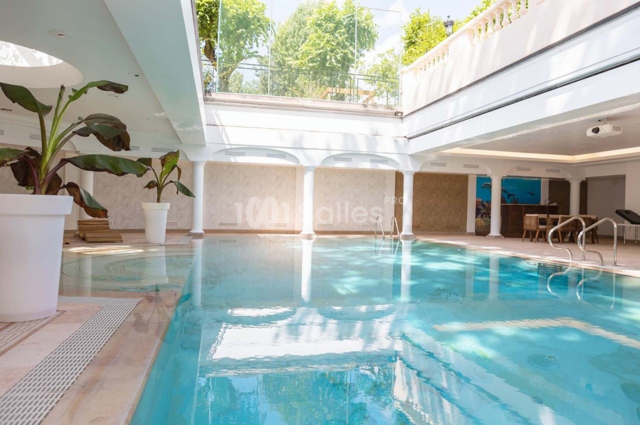 Piscine intérieure lumineuse avec baies vitrées, plantes en pot et mobilier en arrière-plan.