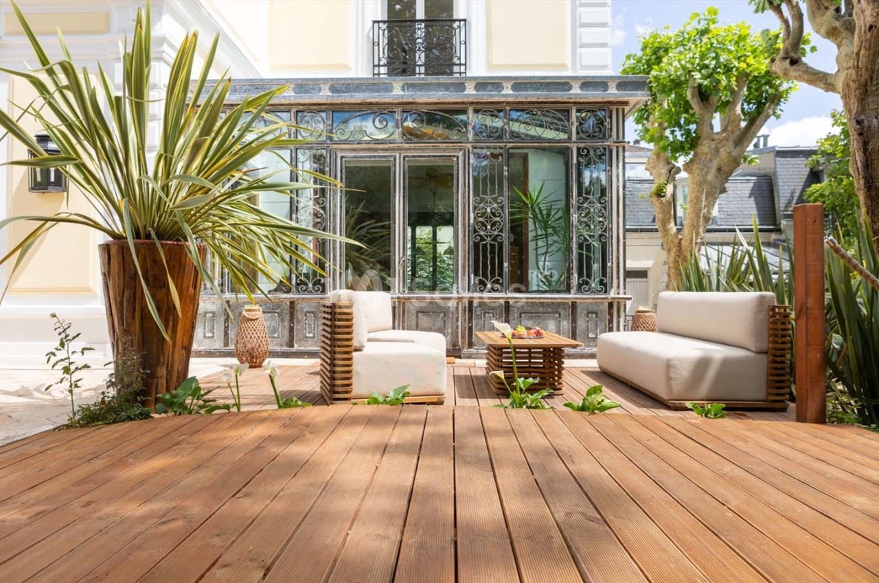 Terrasse en bois avec mobilier de jardin moderne, entourée de plantes et donnant sur une véranda vitrée.