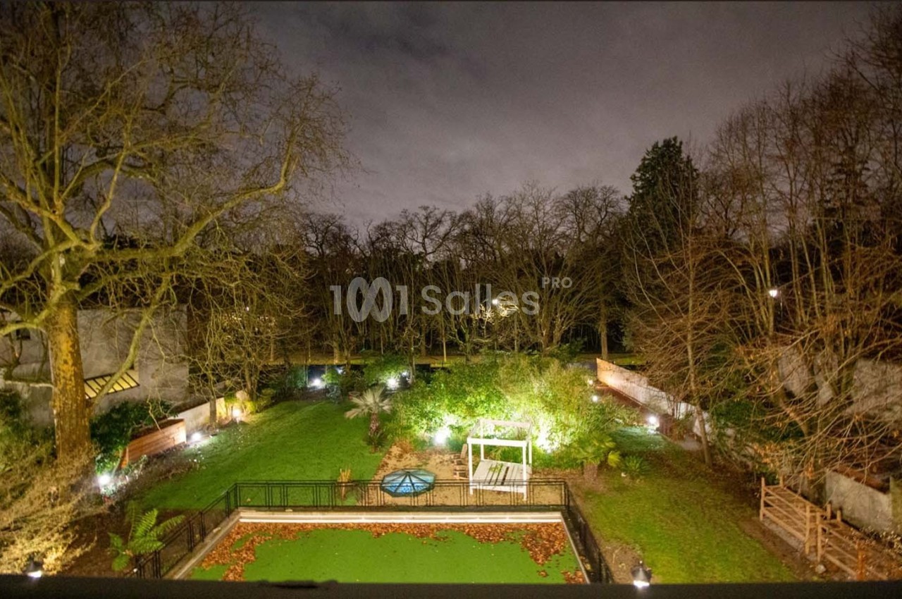 Vue nocturne d'un jardin éclairé avec pelouse, arbres dénudés, bassin central et clôtures en arrière-plan.