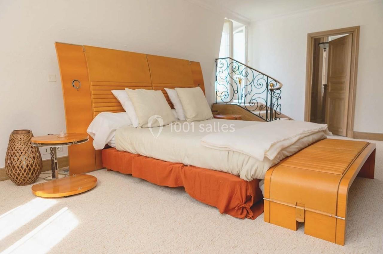 Lit double avec tête de lit en bois, draps clairs, tables de chevet assorties et escalier en fer forgé en arrière-plan.