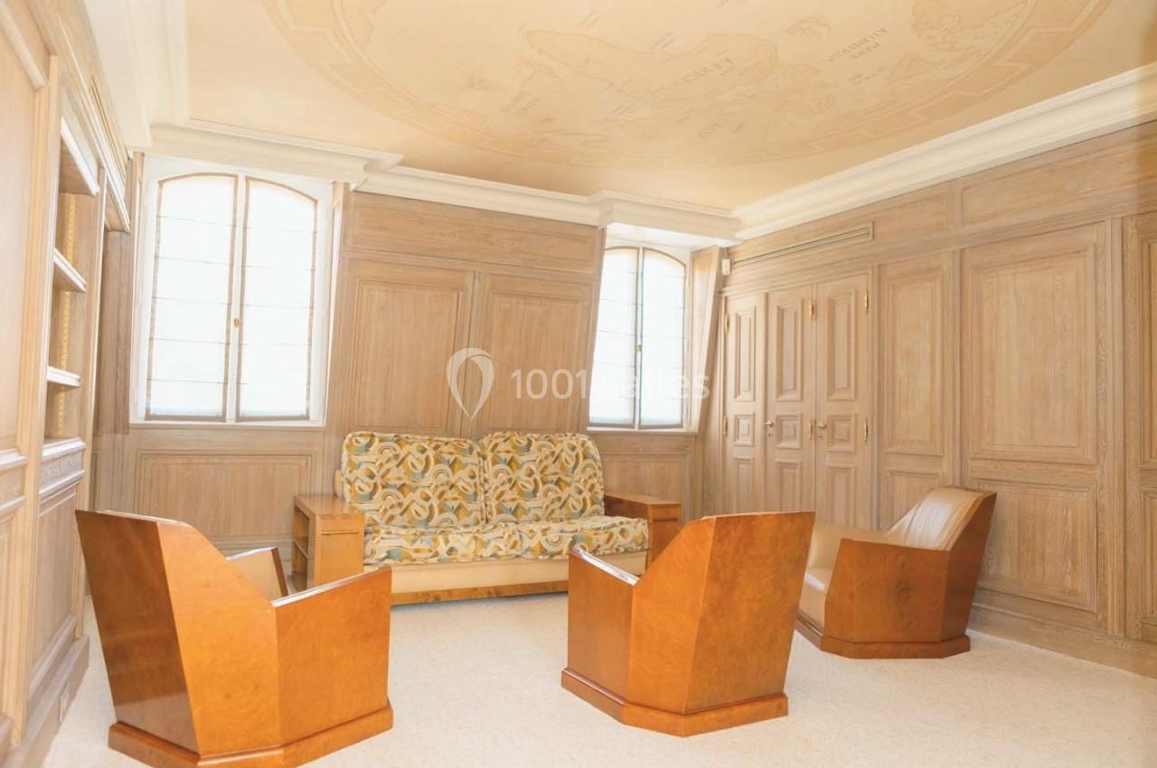 Salle lumineuse avec boiseries murales, canapé à motifs et fauteuils en bois, sous un plafond décoré.