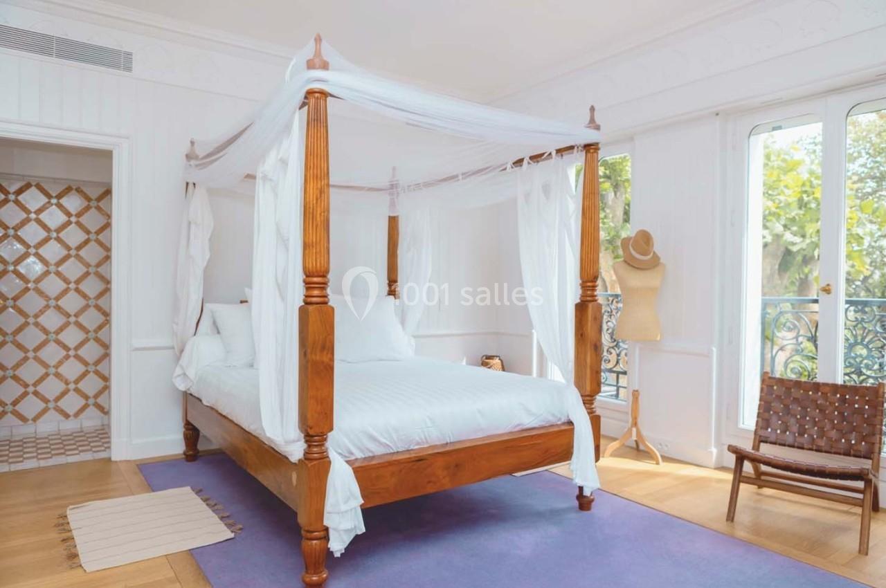 Lit à baldaquin en bois avec voilages blancs dans une chambre lumineuse, parquet au sol et vue sur un balcon.