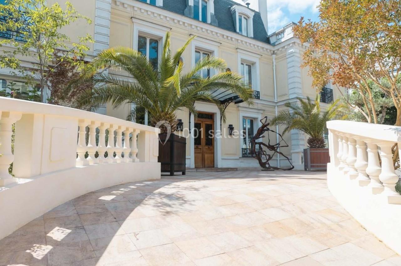 Façade d'un bâtiment élégant avec des palmiers, une sculpture moderne et une terrasse en pierre claire.