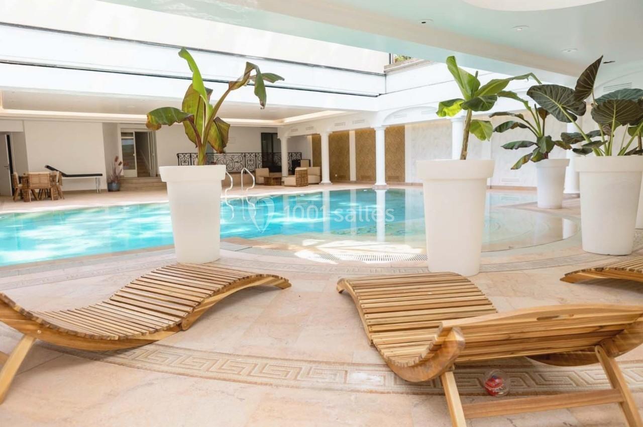 Piscine intérieure entourée de transats en bois et de grandes plantes en pots blancs, dans un espace lumineux.