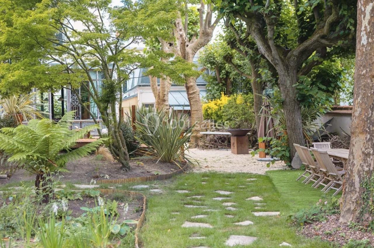 Jardin aménagé avec pelouse, arbres, plantes variées, chemin en pierre et coin repas extérieur avec table et chaises.
