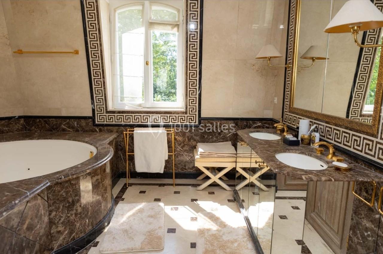 Salle de bain luxueuse avec baignoire en marbre, double vasque, miroirs, et fenêtre donnant sur un jardin.